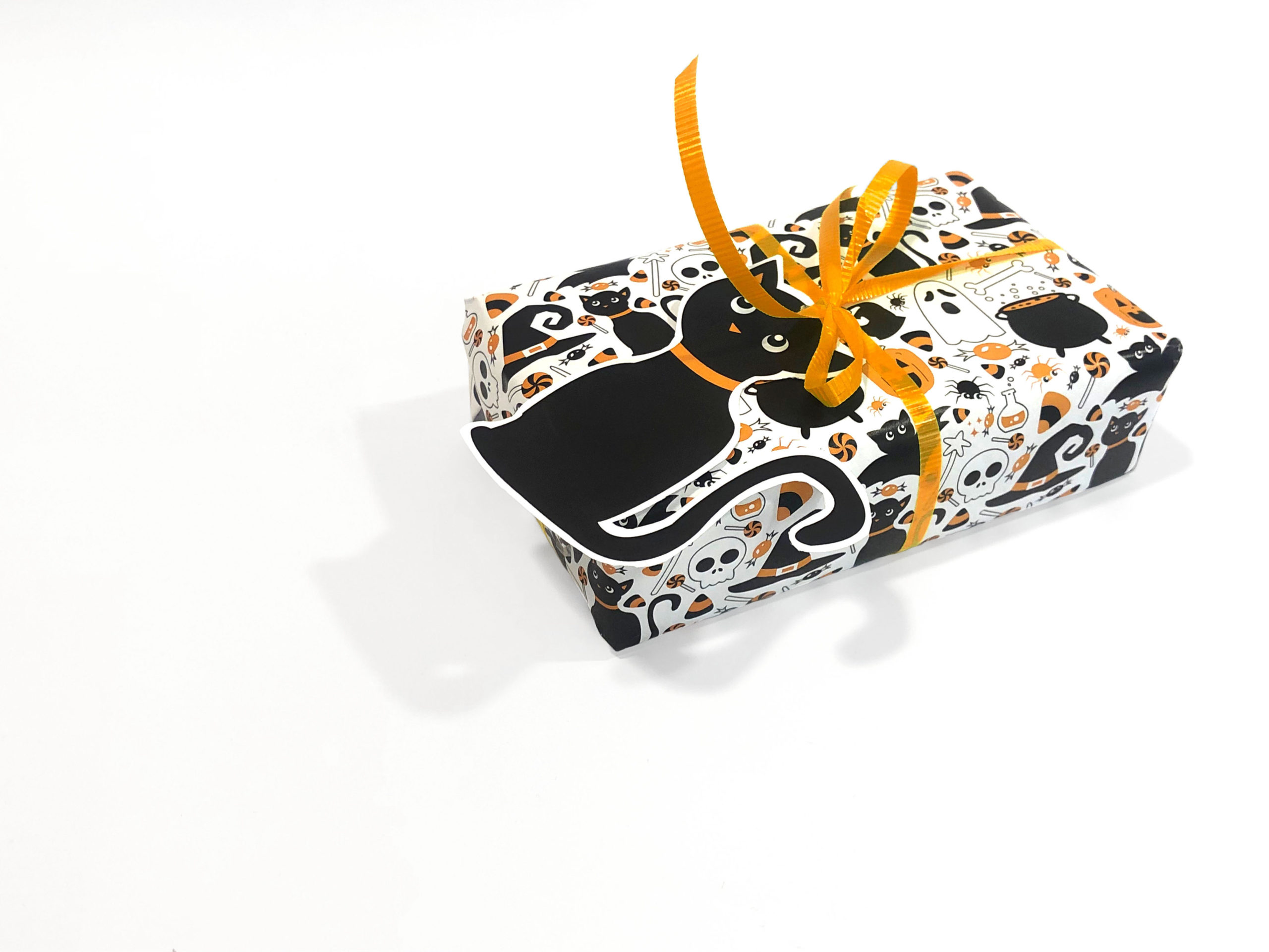 A present wrapped in a pattern with ghosts, jack-o-lanterns, black cats, cauldrons, candy corn, lollipops, caramel apples, wrapped candies, spiders, bats, potion bottles, ghosts, and witches hats. All the objects are are black, orange, and/or white and outlined in black. The background is white. Tied to the present is an orange ribbon with a black cat tag.